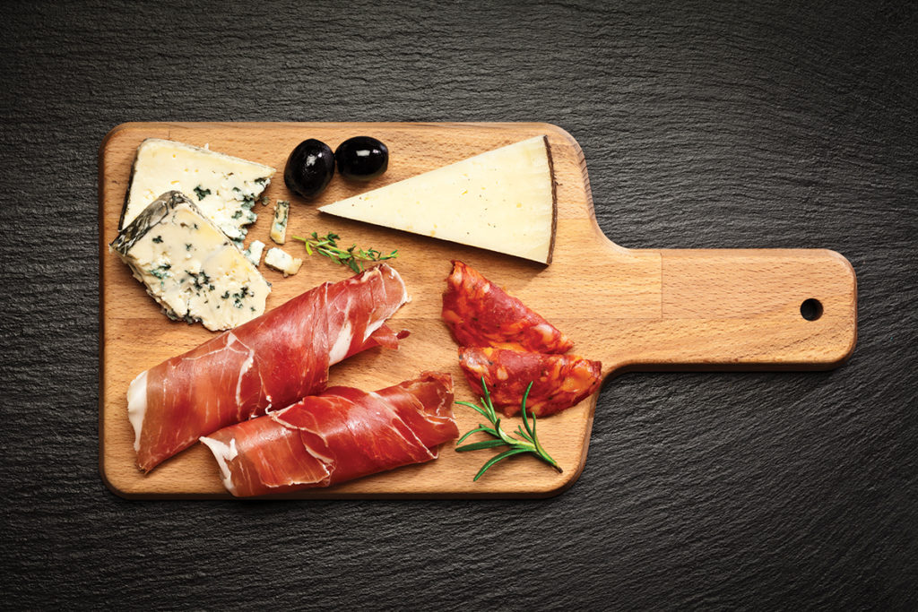 Top view of a wooden cutting board on dark slate background filled with some cold cuts and cheese. DSRL studio photo taken with Canon EOS 5D Mk II and Canon EF 100mm f/2.8L Macro IS USM