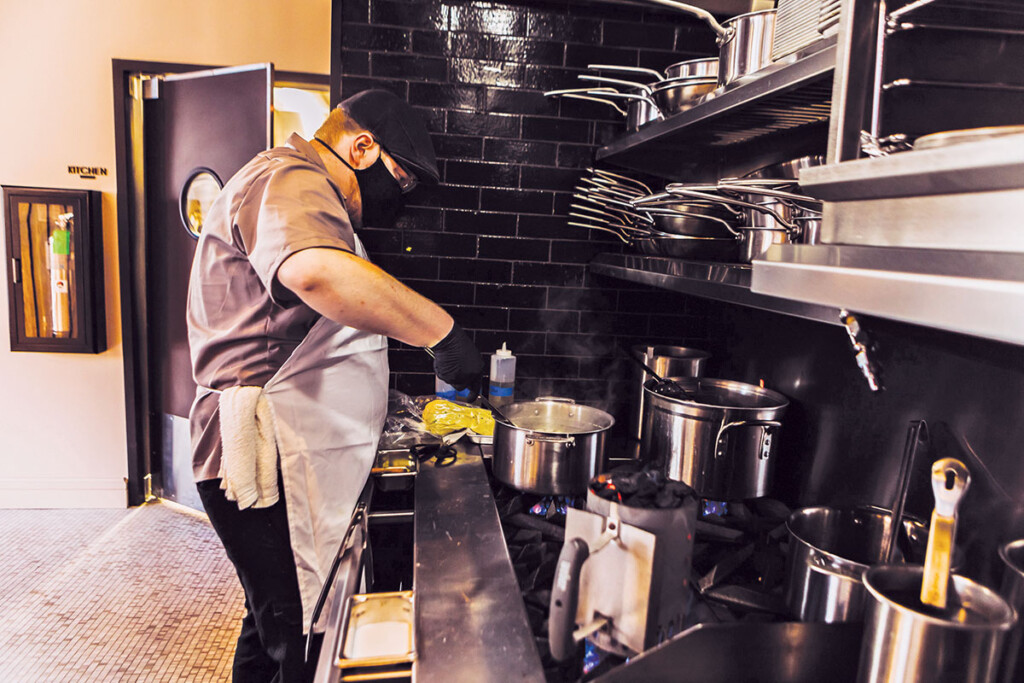 The cookline starts with a range and griddle topped by a cheesemelter. The wood-fired charbroiler occupies the center spot, and a fryer and convection oven complete the line.